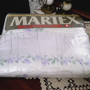 May include: A twin flat sheet made of 100% cotton flannel by Martex, packaged in clear plastic. The sheet is white with a purple floral pattern, a small dot print, and a white lace trim detail along the edge of the fabric design.