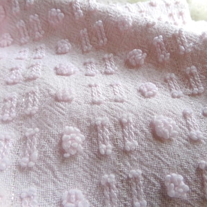 Puede incluir: Primer plano de una tela rosa claro con un patrón texturizado. La tela presenta detalles en relieve, afelpados, en varias formas, incluyendo pequeños círculos y rectángulos alargados. El fondo muestra una sutil textura tejida.