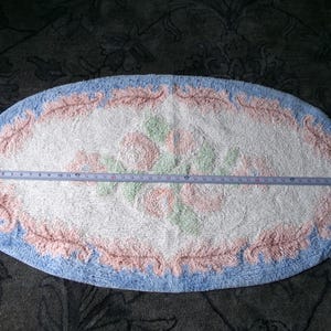 May include: Oval shaped bath mat with a white background and a pink and green floral design. The mat has a blue border with a pink and white floral pattern.
