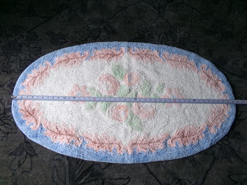 May include: Oval shaped bath mat with a white background and a pink and green floral design. The mat has a blue border with a pink and white floral pattern.