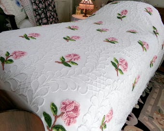 May include: A white bedspread featuring an intricate raised quilted pattern of swirling leaves and vines. The fabric is decorated with evenly spaced embroidered pink rose blossoms with green stems and leaves scattered across the entire surface.