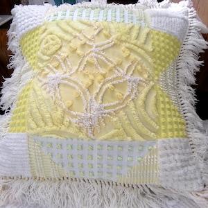 May include: A decorative square pillow with a patchwork design. The pillow features a central yellow panel with white embroidered floral patterns. White and yellow textured fabrics and fringe trim the edges. The pillow is resting on a white surface.