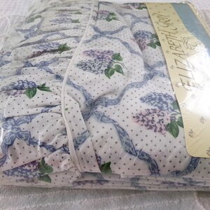 May include: White bed sheet with a blue and purple floral pattern and white polka dots. The sheet has a ruffled edge and is packaged in plastic with the text "Elizabeth Gray" printed on the packaging.