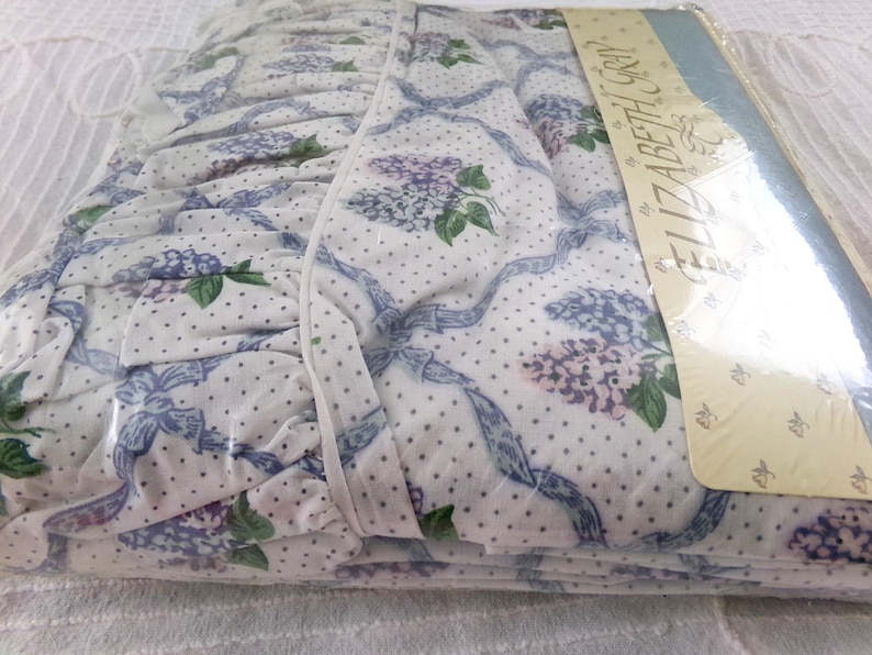 May include: White bed sheet with a blue and purple floral pattern and white polka dots. The sheet has a ruffled edge and is packaged in plastic with the text "Elizabeth Gray" printed on the packaging.