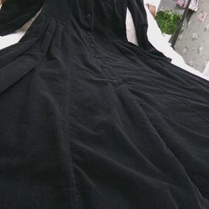 May include: A long-sleeved black dress featuring a button-down front closure, a high neckline, and a flared skirt with vertical seam detailing. The garment is crafted from a textured fabric with a subtle corduroy-like appearance and long sleeves.