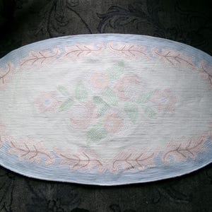 May include: Oval-shaped rug with a light blue border and a white center featuring a floral design in pink and green.