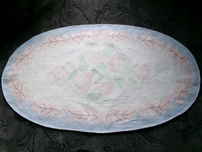 May include: Oval-shaped rug with a light blue border and a white center featuring a floral design in pink and green.