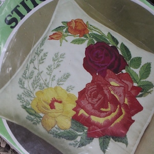May include: A close-up of a white pillow with embroidered roses in shades of red, yellow, and purple. The pillow is packaged in plastic and labeled "Stitchery" with a green background.