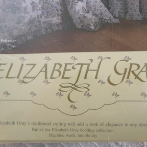 May include: A close-up of a beige label with the text "ELIZABETH GRAY" in gold lettering. The label has a floral design with small pink flowers. The text below the logo reads "Elizabeth Gray's traditional styling will add a look of elegance to any decor. Part of the Elizabeth Gray bedding collection. Machine wash, tumble dry."