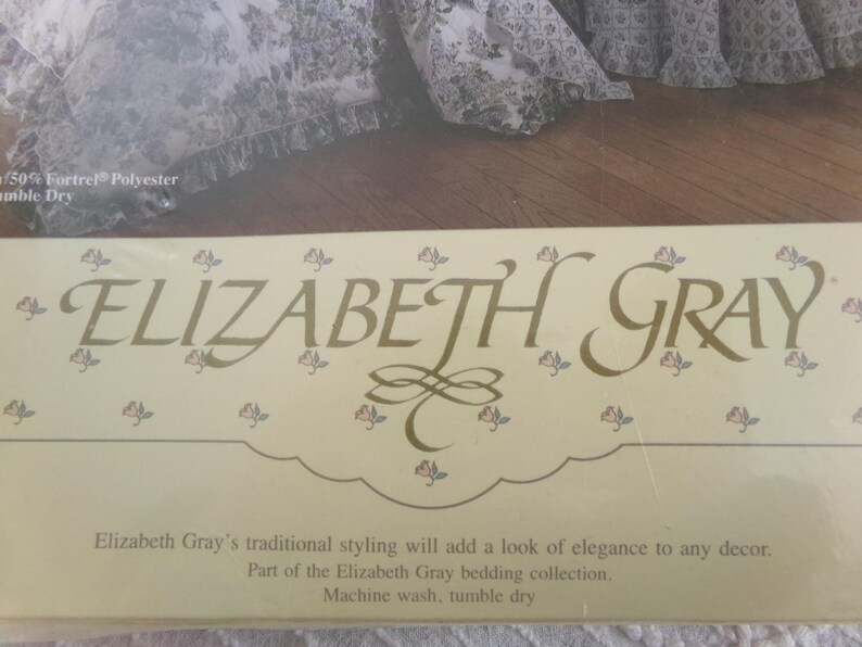 May include: A close-up of a beige label with the text "ELIZABETH GRAY" in gold lettering. The label has a floral design with small pink flowers. The text below the logo reads "Elizabeth Gray's traditional styling will add a look of elegance to any decor. Part of the Elizabeth Gray bedding collection. Machine wash, tumble dry."