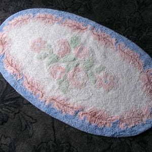 May include: Oval shaped bath mat with a pink and blue floral design. The mat is white in the center with a pink and blue border.