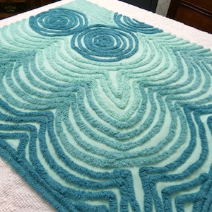 May include: A diamond-shaped, tufted rug in shades of teal and turquoise. The rug features a repeating pattern of concentric circles and curved lines, creating a textured, abstract design. The rug's edges are outlined with a darker teal, adding definition to the overall shape.