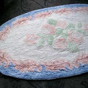 May include: A white oval bath mat with a blue and pink border. The center of the mat features a floral design with pink, green, and white flowers.