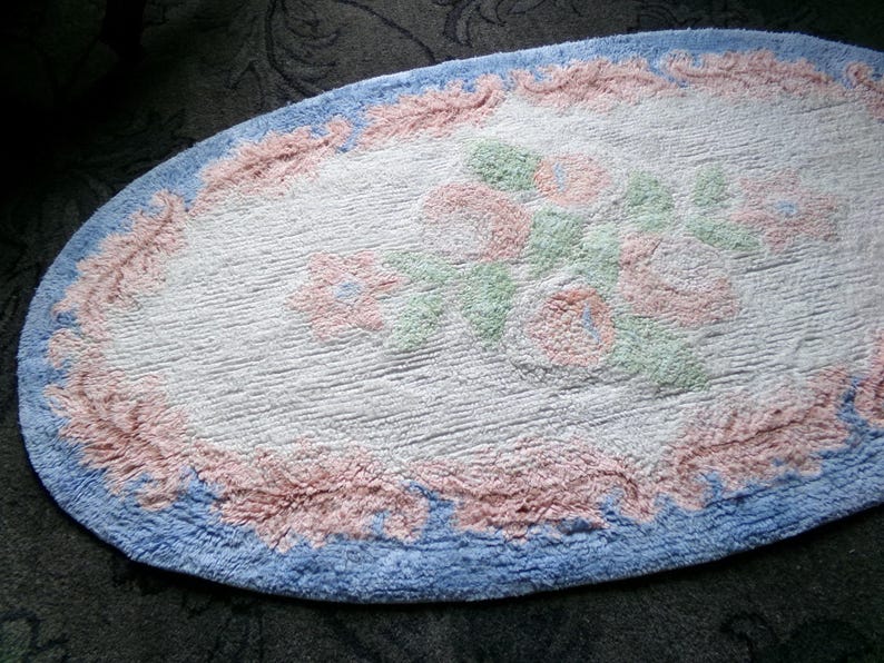 May include: A white oval bath mat with a blue and pink border. The center of the mat features a floral design with pink, green, and white flowers.