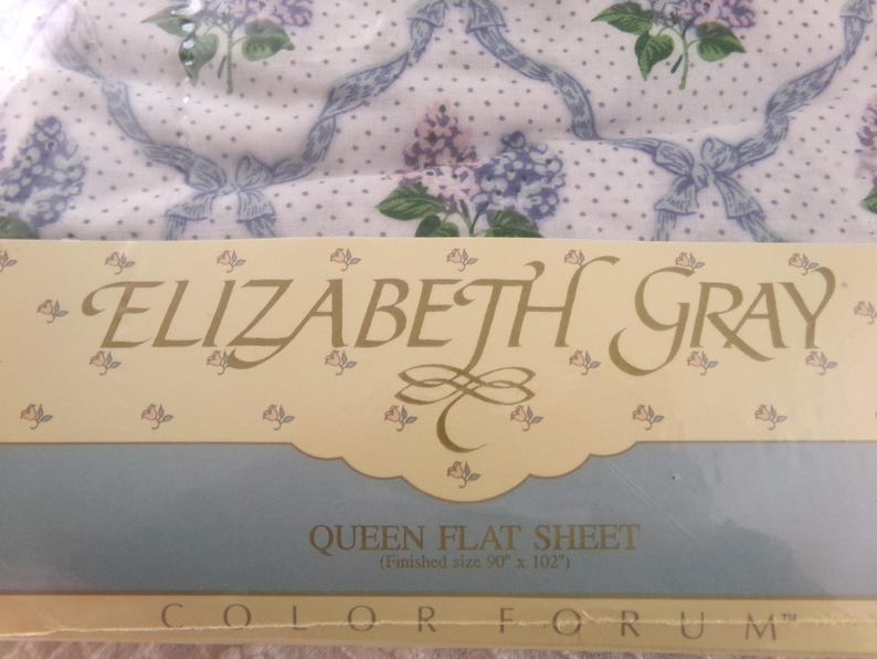 May include: A white flat sheet with a floral pattern in shades of purple and green. The sheet is labeled "ELIZABETH GRAY QUEEN FLAT SHEET (Finished size 90 x 102") COLOR FORUM".