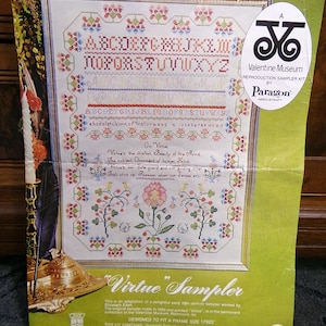 May include: A vintage cross-stitch sampler kit titled "Virtue Sampler." The kit features a framed design with floral and alphabet patterns in red, green, and blue. The kit includes stamped linen, embroidery thread, and instructions. The frame size is 17x23 inches.