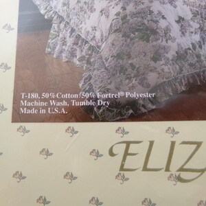 May include: A white and purple floral bed skirt with a ruffled edge. The fabric is 50% cotton and 50% polyester. The text "T-180, 50% Cotton/50% Fortrel&reg; Polyester Machine Wash, Tumble Dry Made in U.S.A." is printed on the packaging. The text "ELIZA" is printed on the bottom of the packaging.