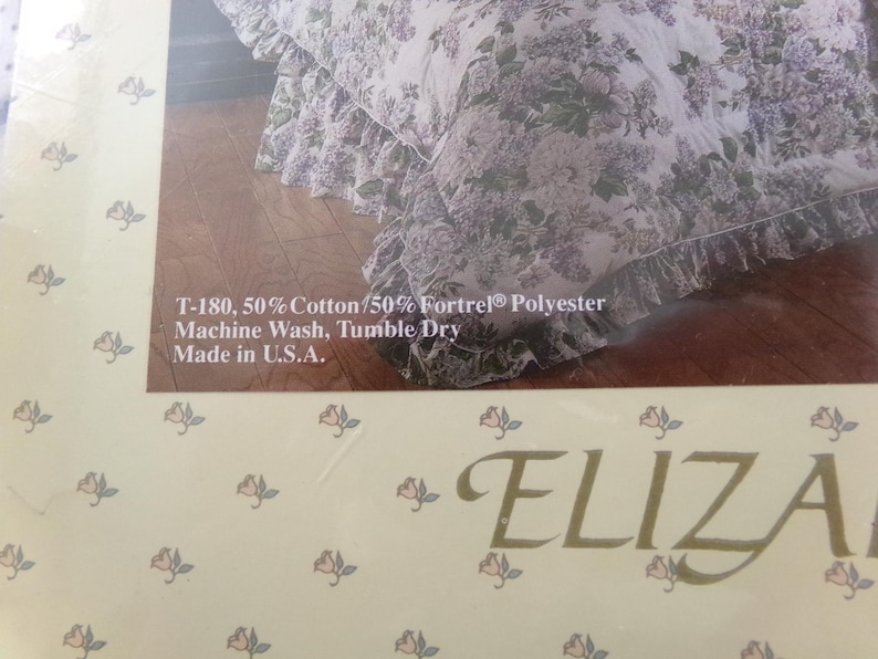 May include: A white and purple floral bed skirt with a ruffled edge. The fabric is 50% cotton and 50% polyester. The text "T-180, 50% Cotton/50% Fortrel&reg; Polyester Machine Wash, Tumble Dry Made in U.S.A." is printed on the packaging. The text "ELIZA" is printed on the bottom of the packaging.