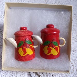May include: Two small, red wooden teapots with white spouts and handles, decorated with a cherry design. The teapots are inside a beige box with white padding. The teapots are likely decorative items or toys.
