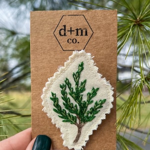 Puede incluir: Un pequeño árbol verde cosido a mano bordado en un parche de tela blanca. El parche está unido a una tarjeta marrón con el texto "d+m co."