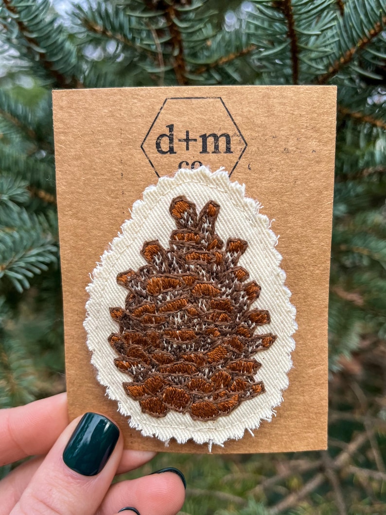 May include: A brown embroidered pine cone patch on a white fabric background with a scalloped edge. The patch is on a brown card with the text "d+m co" in black.