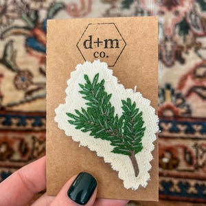 K&ouml;nnte beinhalten: Ein gr&uuml;ner gestickter Kiefernzweig auf einem wei&szlig;en Stoffst&uuml;ck mit braunem Papieruntergrund. Das St&uuml;ck hat einen ausgefransten Rand und der Text "d+m co." ist auf den Untergrund gedruckt.