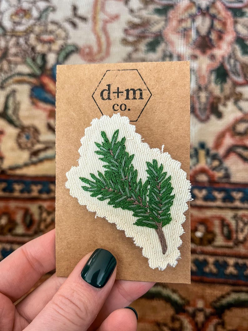 K&ouml;nnte beinhalten: Ein gr&uuml;ner gestickter Kiefernzweig auf einem wei&szlig;en Stoffst&uuml;ck mit braunem Papieruntergrund. Das St&uuml;ck hat einen ausgefransten Rand und der Text "d+m co." ist auf den Untergrund gedruckt.