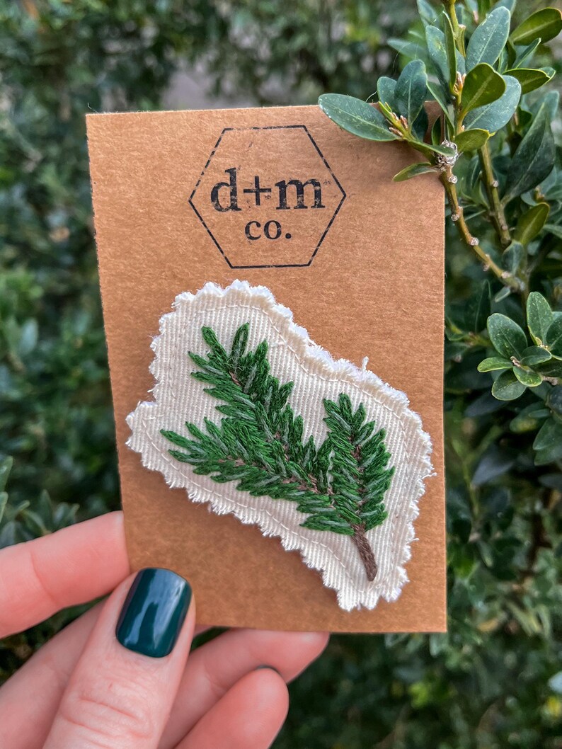 K&ouml;nnte beinhalten: Ein gr&uuml;ner gestickter Kiefernzweig auf einem wei&szlig;en Stoffst&uuml;ck mit braunem Papierhintergrund. Das St&uuml;ck hat einen gewellten Rand. Der Papierhintergrund hat ein Sechseck mit dem Text "d+m co."