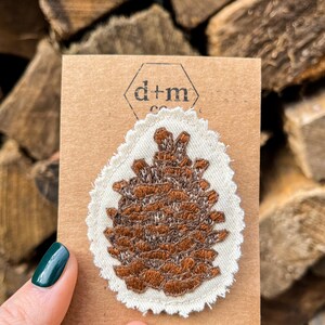 May include: A brown embroidered pine cone patch on a white fabric background with a scalloped edge. The patch is on a brown card with the text "d+m co."