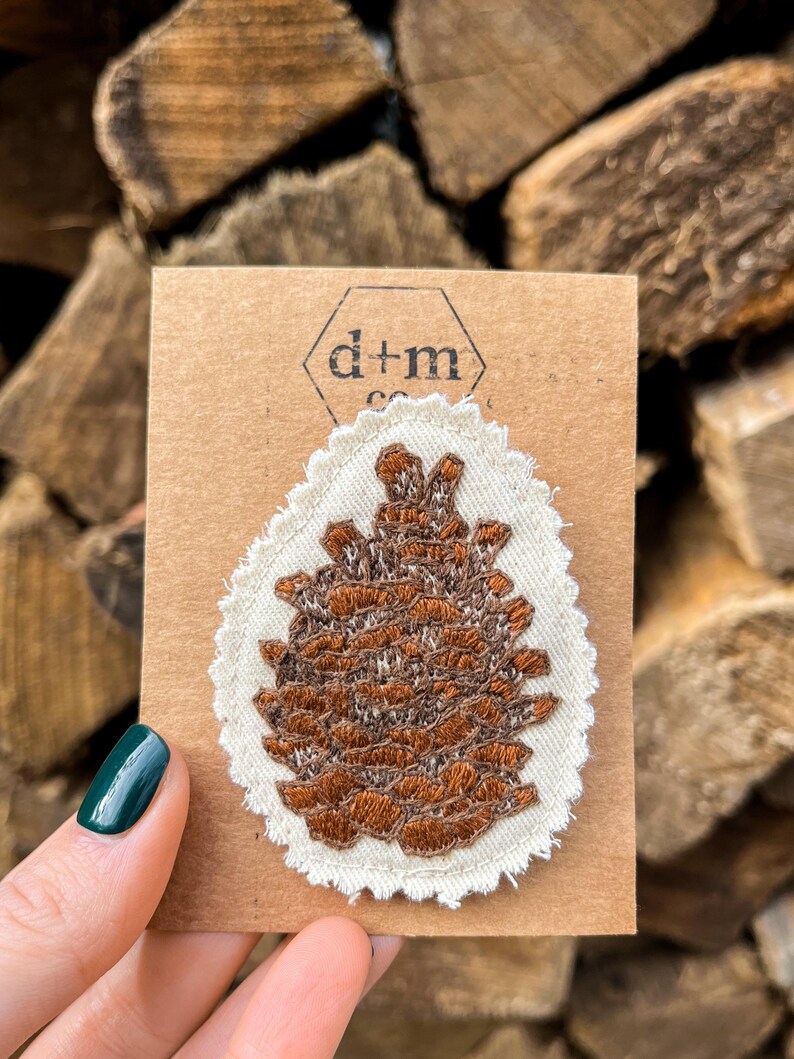 May include: A brown embroidered pine cone patch on a white fabric background with a scalloped edge. The patch is on a brown card with the text "d+m co."