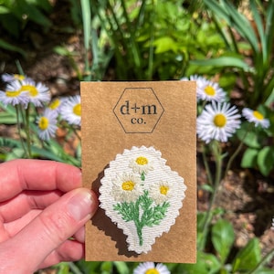 Op de afbeelding: Een witte stoffen patch met een geborduurd ontwerp van drie witte madeliefjes met groene stelen en bladeren. De patch is bevestigd aan een bruine kaart met de tekst "d+m co."