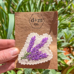 May include: A white fabric patch with purple embroidered lavender flowers. The patch is attached to a brown card with the text "d+m co."