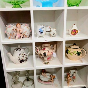 Puede incluir: Exhibici&oacute;n de tazas de t&eacute;, teteras y objetos decorativos vintage en una estanter&iacute;a blanca. Los art&iacute;culos incluyen dise&ntilde;os florales, figuras de querubines y cuencos de vidrio. La paleta de colores es suave, con pasteles y blancos.