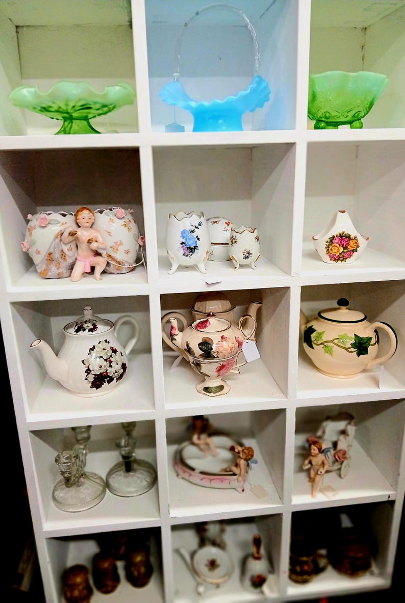 Puede incluir: Exhibici&oacute;n de tazas de t&eacute;, teteras y objetos decorativos vintage en una estanter&iacute;a blanca. Los art&iacute;culos incluyen dise&ntilde;os florales, figuras de querubines y cuencos de vidrio. La paleta de colores es suave, con pasteles y blancos.