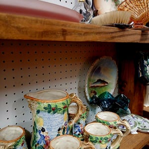 Puede incluir: Una colecci&oacute;n de jarras y tazas de cer&aacute;mica vintage con un dise&ntilde;o de castillo. La jarra azul tiene un asa detallada y una escena de personas. Las tazas tienen un dise&ntilde;o similar con detalles verdes y rojos. Otros art&iacute;culos vintage est&aacute;n en los estantes.