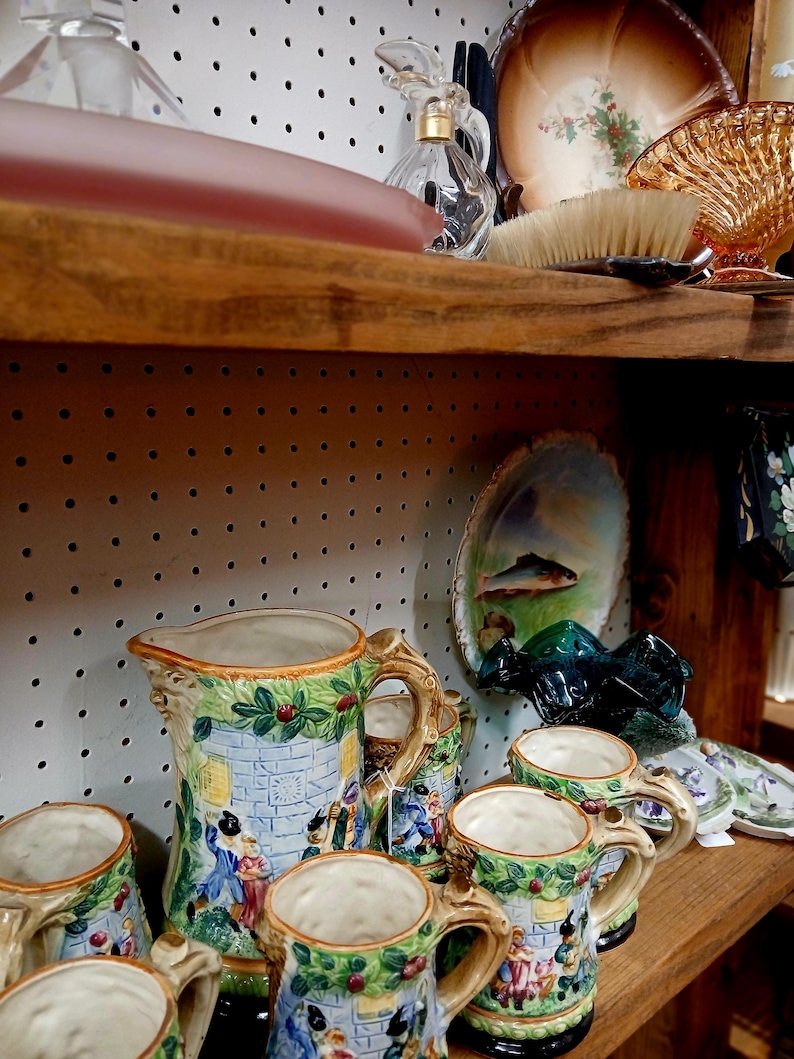 Puede incluir: Una colecci&oacute;n de jarras y tazas de cer&aacute;mica vintage con un dise&ntilde;o de castillo. La jarra azul tiene un asa detallada y una escena de personas. Las tazas tienen un dise&ntilde;o similar con detalles verdes y rojos. Otros art&iacute;culos vintage est&aacute;n en los estantes.