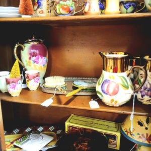 Puede incluir: Una estanter&iacute;a de madera exhibe tazas de t&eacute;, teteras y objetos decorativos antiguos. Las tazas de t&eacute; y las teteras presentan dise&ntilde;os florales y de frutas. Tambi&eacute;n se exhiben un peque&ntilde;o &aacute;rbol en forma de cono naranja y un &aacute;rbol dorado.