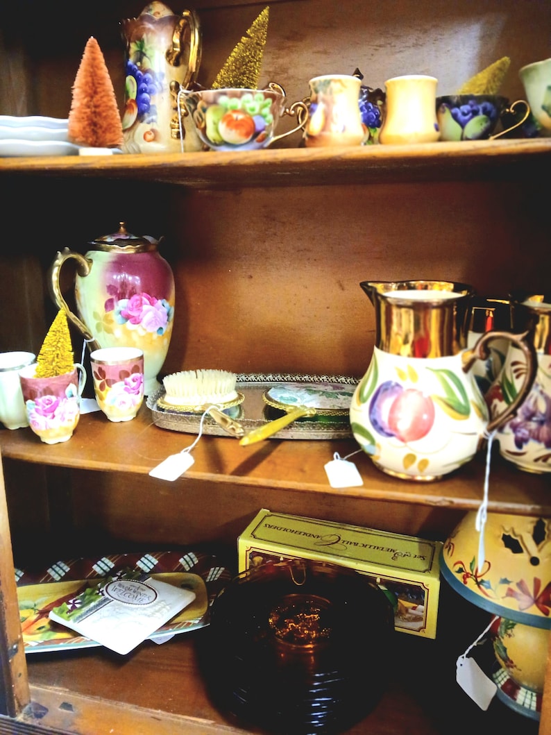 Puede incluir: Una estanter&iacute;a de madera exhibe tazas de t&eacute;, teteras y objetos decorativos antiguos. Las tazas de t&eacute; y las teteras presentan dise&ntilde;os florales y de frutas. Tambi&eacute;n se exhiben un peque&ntilde;o &aacute;rbol en forma de cono naranja y un &aacute;rbol dorado.