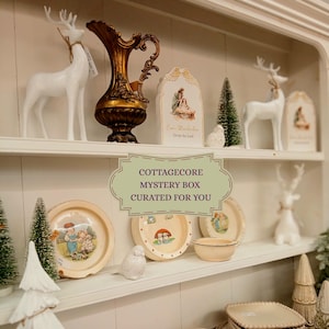 Puede incluir: Una exhibici&oacute;n de decoraci&oacute;n cottagecore en estantes blancos. Los art&iacute;culos incluyen figuras de ciervos blancos, platos decorativos con ilustraciones, un jarr&oacute;n dorado, peque&ntilde;os &aacute;rboles de Navidad y un letrero que dice "COTTAGECORE MYSTERY BOX CURATED FOR YOU."