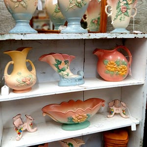 Puede incluir: Una colecci&oacute;n de jarrones de cer&aacute;mica vintage y piezas decorativas en colores pastel. Los jarrones presentan dise&ntilde;os florales y se exhiben en estantes blancos. La paleta de colores incluye rosa, azul, amarillo y melocot&oacute;n.