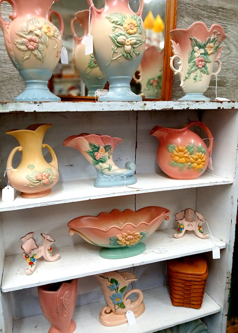 Puede incluir: Una colecci&oacute;n de jarrones de cer&aacute;mica vintage y piezas decorativas en colores pastel. Los jarrones presentan dise&ntilde;os florales y se exhiben en estantes blancos. La paleta de colores incluye rosa, azul, amarillo y melocot&oacute;n.