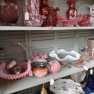 Puede incluir: Una colecci&oacute;n de cristaler&iacute;a y objetos decorativos vintage exhibidos en estantes. Los art&iacute;culos incluyen jarrones, cuencos y una cesta en tonos rosa, rojo y vidrio transparente. Algunas piezas tienen bordes con volantes o dise&ntilde;os intrincados.