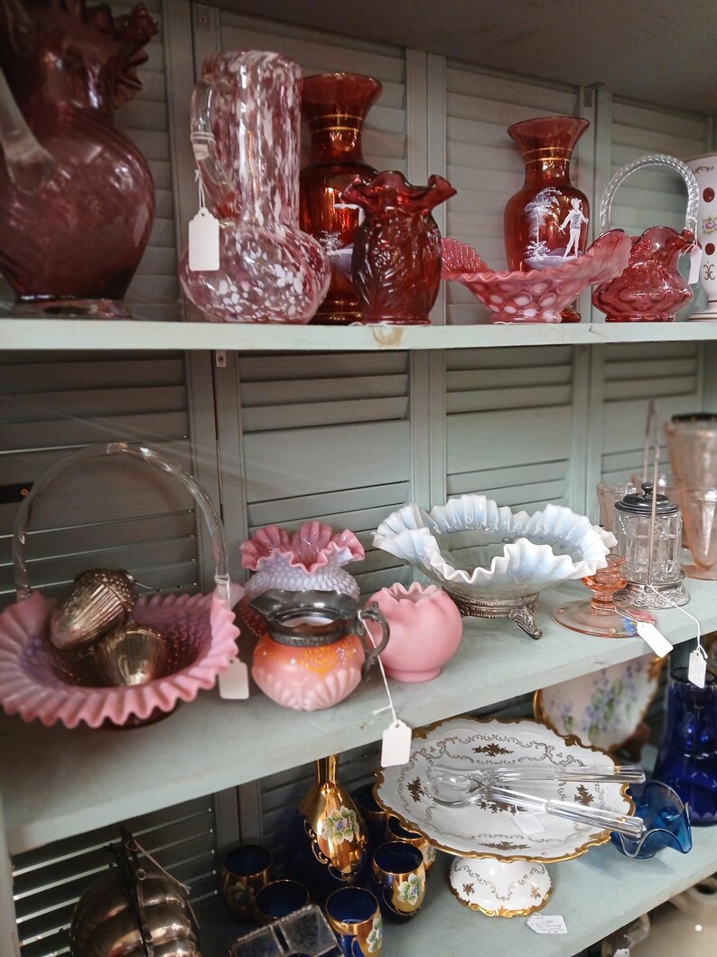 Puede incluir: Una colecci&oacute;n de cristaler&iacute;a y objetos decorativos vintage exhibidos en estantes. Los art&iacute;culos incluyen jarrones, cuencos y una cesta en tonos rosa, rojo y vidrio transparente. Algunas piezas tienen bordes con volantes o dise&ntilde;os intrincados.