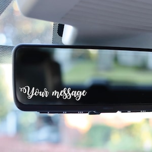 May include: A black car rearview mirror with the words "Your message" in white script. The mirror is mounted inside a vehicle, with a blurred view of the outside visible through the glass.