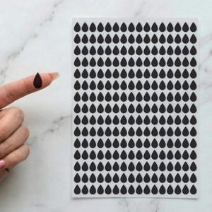 May include: A rectangular sheet of white adhesive paper featuring a grid of numerous small, solid black teardrop-shaped stickers arranged in uniform rows and columns, with one individual black teardrop sticker held separately to the side.