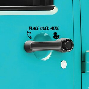 May include: A bright turquoise vehicle door featuring a black horizontal door handle and a black decal above it. The decal includes the text PLACE DUCK HERE, a small black rubber duck icon, and a curved arrow pointing toward the door handle area.