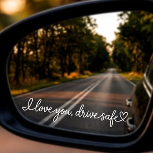 May include: A black car side mirror with a white vinyl decal applied to the glass. The decal features the cursive text "I love you, drive safe" followed by a small heart outline. The mirror reflects a paved road lined with trees at sunset.