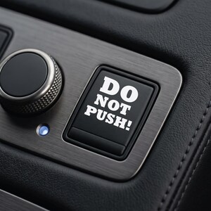May include: A black square push button featuring the white text DO NOT PUSH! in a bold font, set within a brushed metallic panel next to a circular control knob and a small glowing blue indicator light on a dark textured surface.