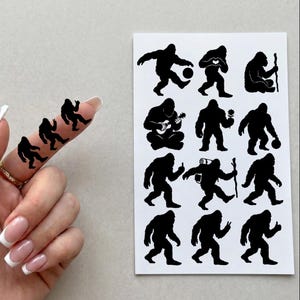 Op de afbeelding: Een vel wit papier met twaalf zwarte silhouetstickers van Bigfoot in verschillende houdingen, waaronder sporten, een bloem vasthouden en wandelen. Drie kleinere bijpassende Bigfoot-silhouetstickers zijn op een vinger aangebracht.