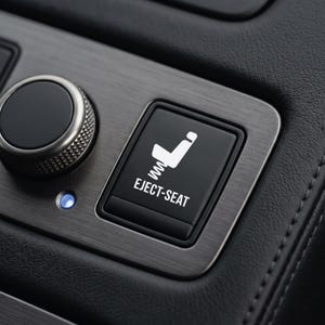May include: A black square push button featuring a white icon of a seat with a spring underneath and the text EJECT-SEAT in white lettering. The button is set into a brushed metallic panel next to a circular dial and a small blue indicator light.